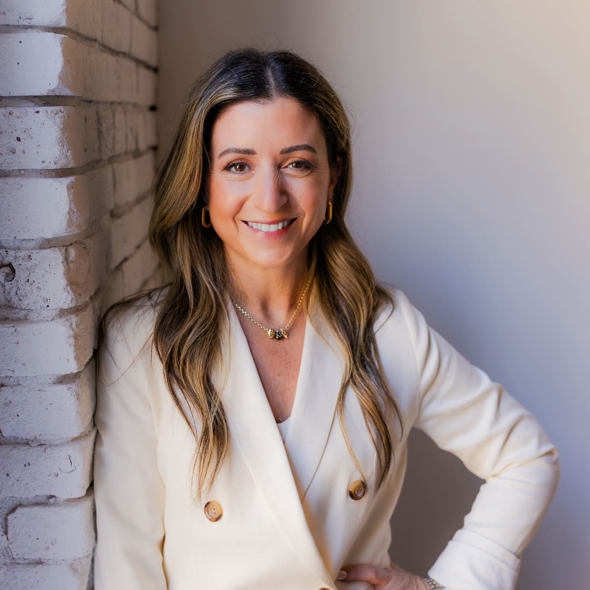 Jackie Giannelli, Board Certified Nurse Practitioner wearing an ivory blazer with gold buttons and her hand on her hip, leaning on a white washed brick wall
