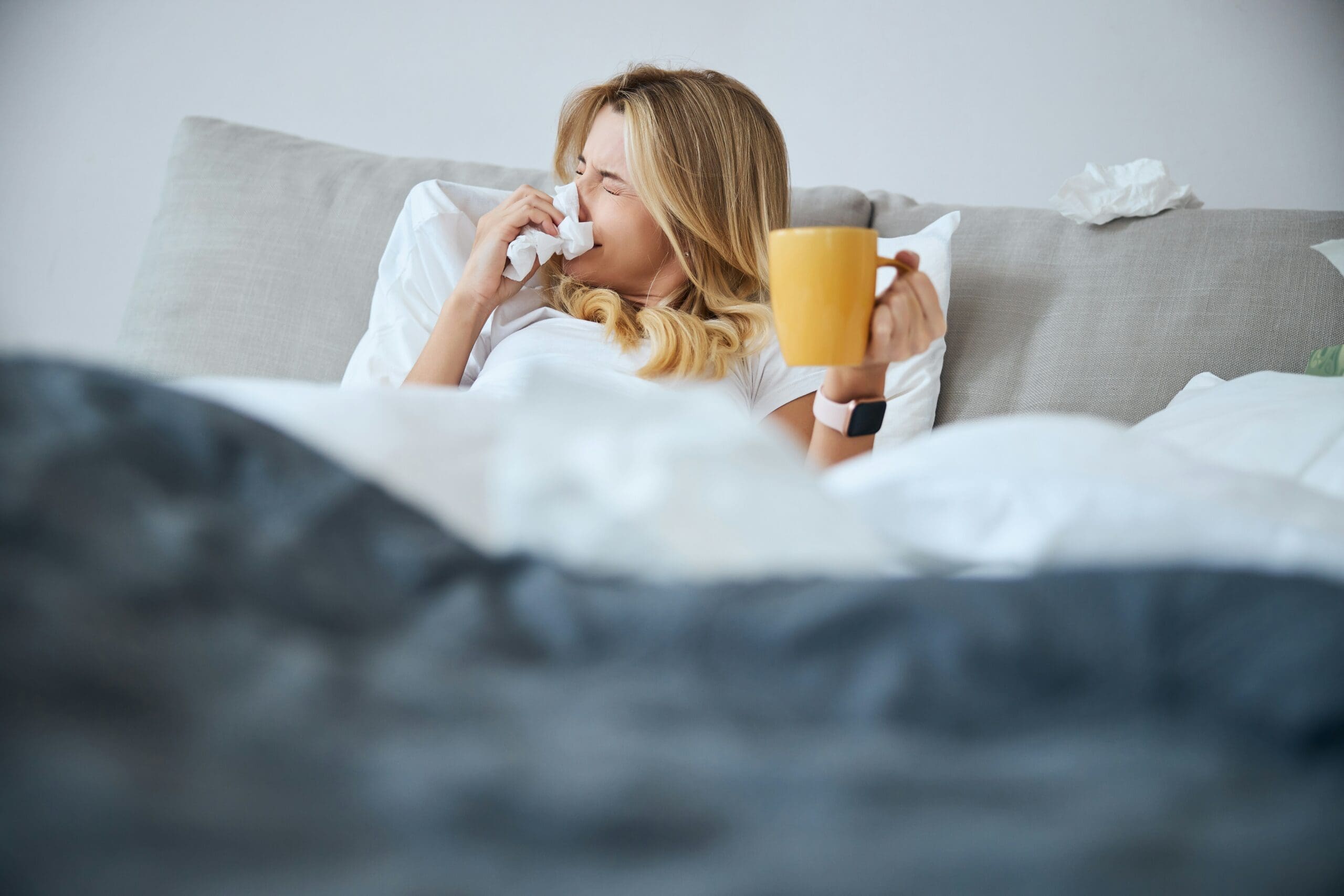 Blonde woman in bed holding a yellow mug and blowing her nose on a kleenex